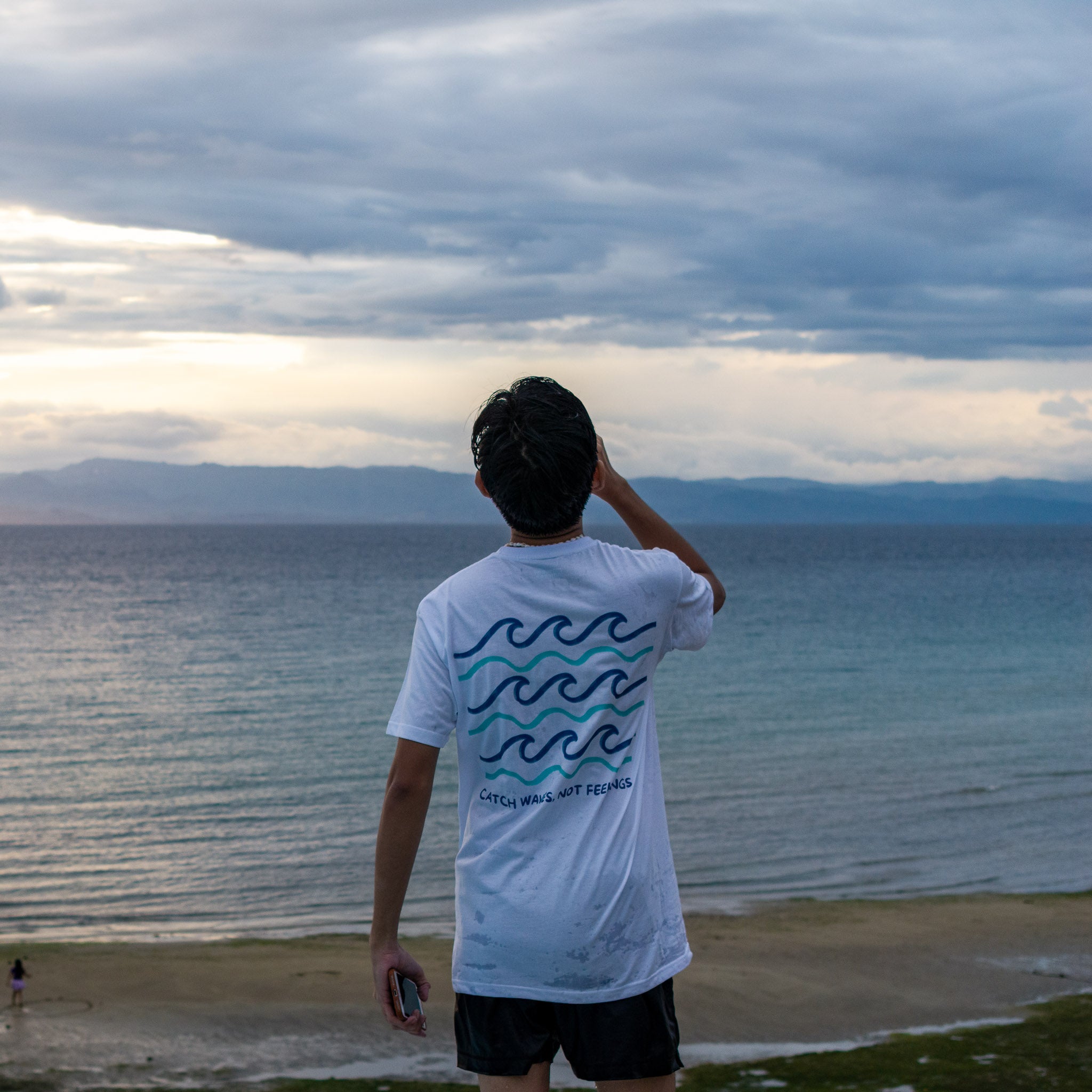 Person wearing a white t-shirt with wave design standing on a beach looking at the ocean.