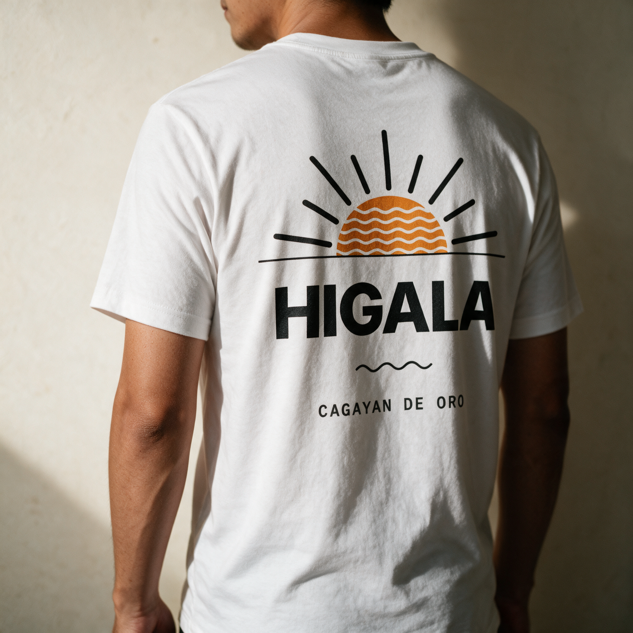 Man seen from behind in the white Higala tee against a pale wall, orange sunrise print and Cagayan de Oro across his back