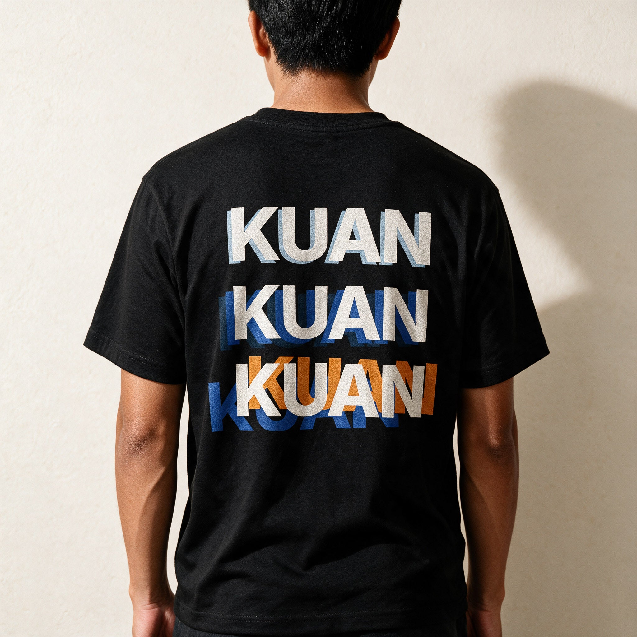 Man seen from behind in the Midnight Black Kuan tee, the word stacked three times across his back in blue and orange