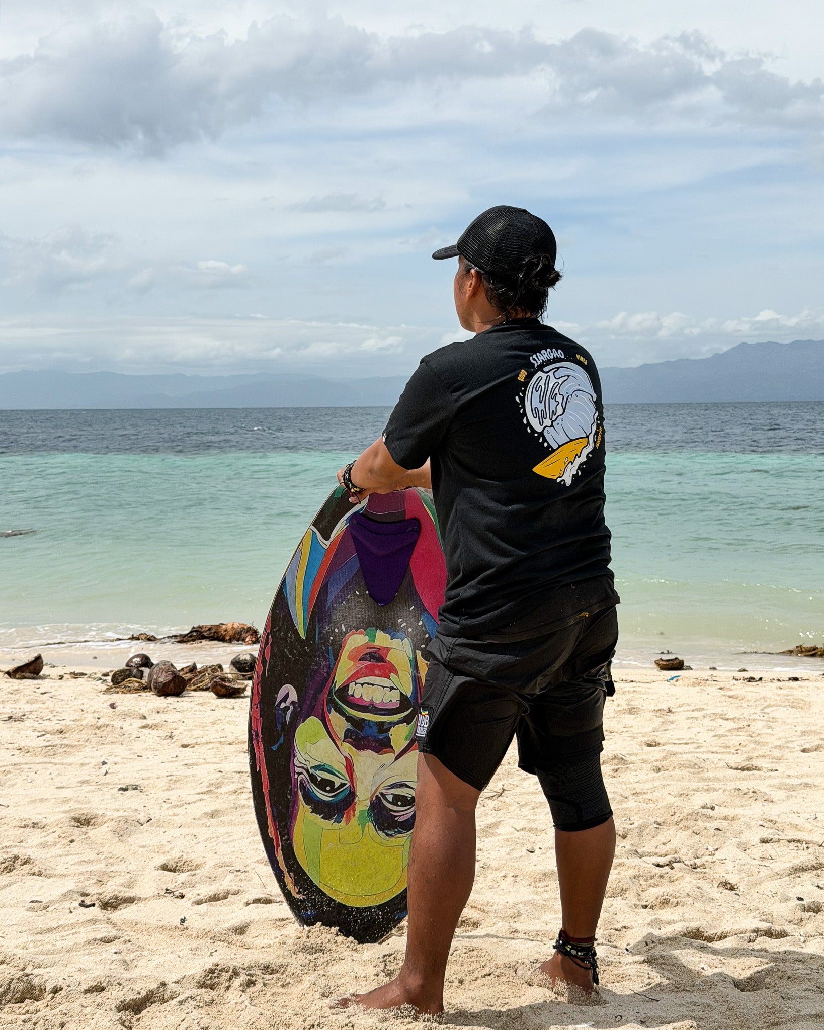 Black Siargao Surf Vibes tee worn by a man holding a skimboard at the shore