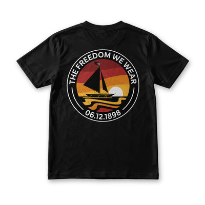 The Freedom We Wear tee in black, back view, paraw badge over a red and gold sunset with 06.12.1898 below