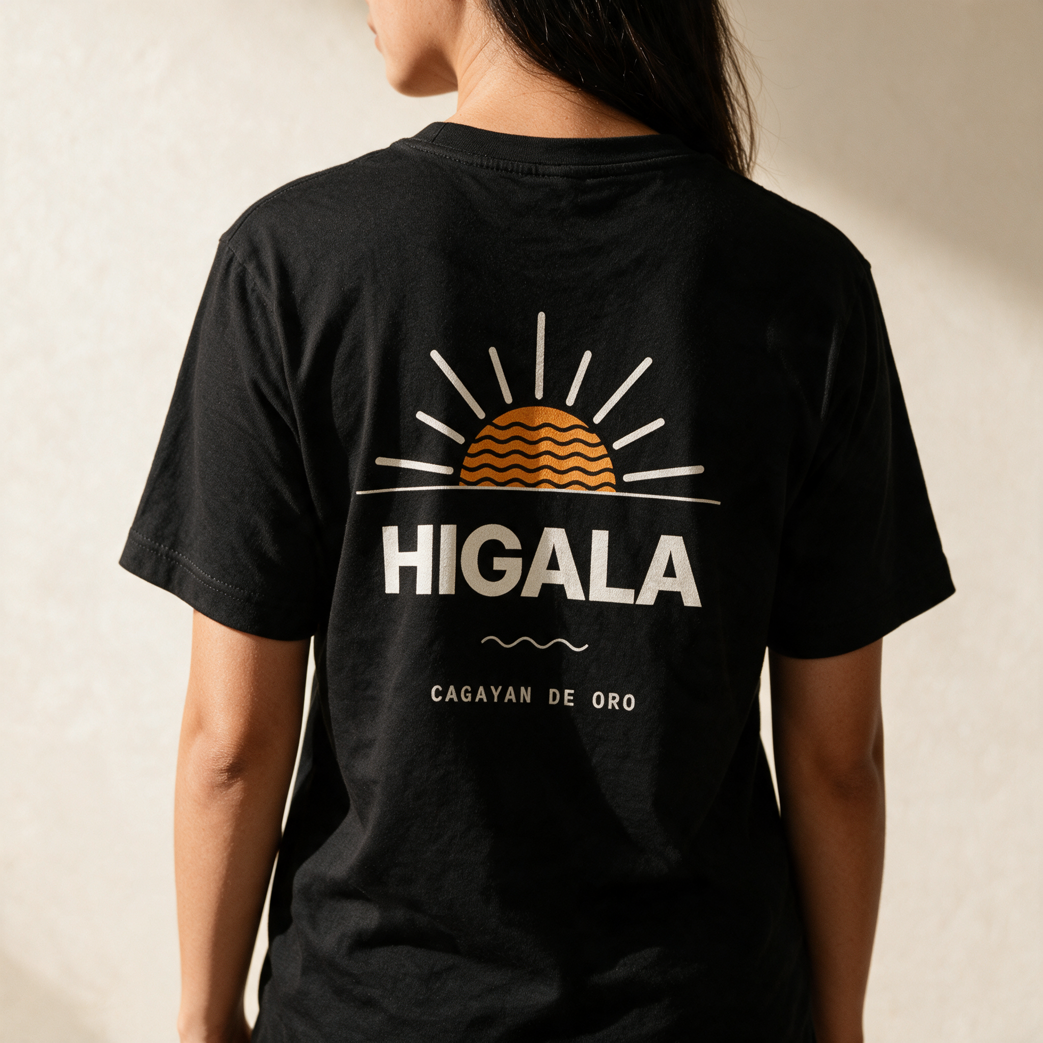 Woman seen from behind in the Midnight Black Higala tee against a pale wall, orange sunrise print and Cagayan de Oro across her back