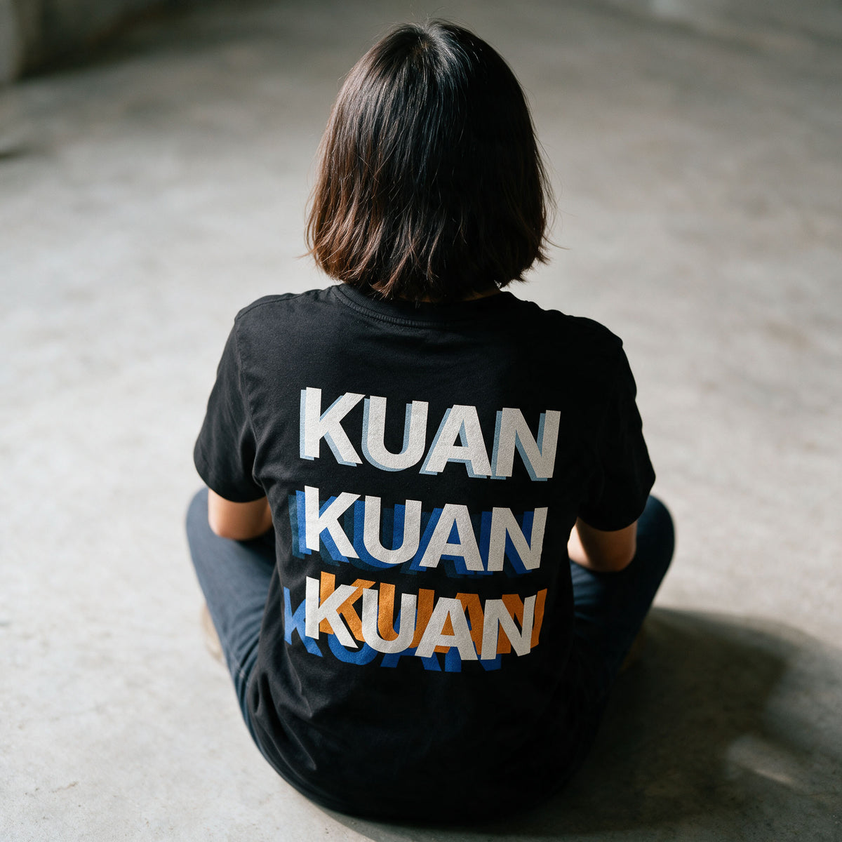 Close-up of the Kuan tee back print in Midnight Black, the word stacked three times out of register in white, blue and orange