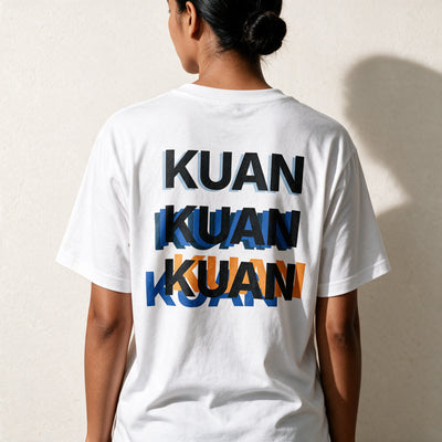 Woman seen from behind in the white Kuan tee, the word stacked three times across her back in blue and orange