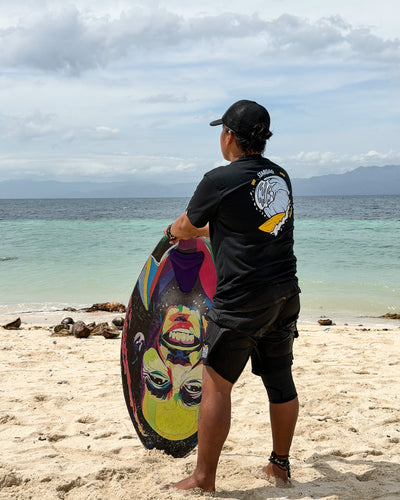 Black Siargao Surf Vibes tee worn by a man holding a skimboard at the shore